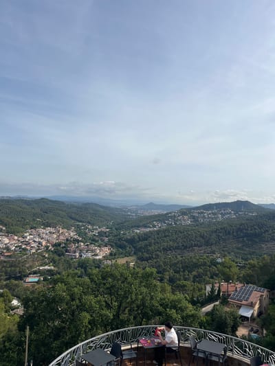 The image depicts an aerial view of a town nestled within a valley surrounded by lush green hills. In the foreground, a person sits at a table on an outdoor balcony, seemingly engaged in a meal or drink. The balcony overlooks a town with terracotta roofs and a sprawling landscape of trees and buildings. In the distance, more hills and a hazy sky with scattered clouds complete the scene. The location is identified as Can Güell, Spain.