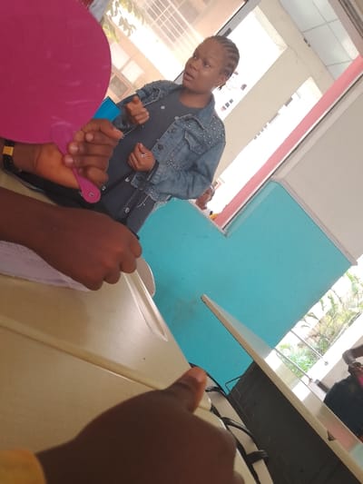 A woman in a studded denim jacket stands inside a building in Umueshi, Nigeria, facing the camera with her mouth slightly open as if speaking. To her right, a window reveals greenery and building features. The interior walls are painted a light turquoise color, contrasted by white trim. In the foreground, the out-of-focus hands of a person holding a pink handheld fan are visible. A desk or table takes up most of the lower part of the frame, partially obscuring the view. Additional people are partially visible in the background.