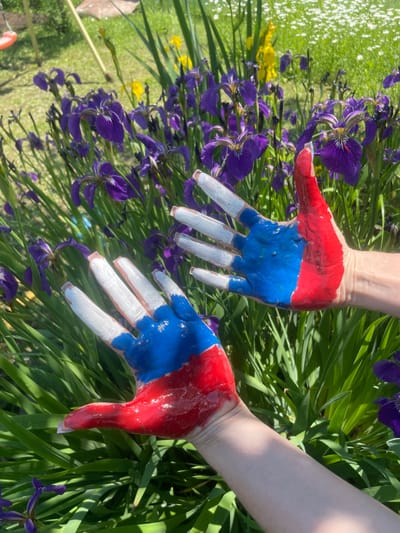 Two human hands are painted with red, white, and blue paint, mimicking the colors of the Russian flag. The hands are positioned in the foreground, with fingers spread apart and palms facing the camera. In the background, a garden setting is visible, featuring purple iris flowers and green foliage. The scene appears to be outdoors on a sunny day. There is no information in the image to suggest it is located in the city of Yasnoe, Russia.