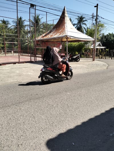 Two individuals, wearing hijabs, ride on a dark-colored scooter. The scooter is moving on an asphalt road. The person in the front is wearing a black hijab and a dark top. The person in the back is wearing a light brown hijab and an orange skirt. The scooter has red and black accents. In the background, there is a fenced sports court, a white tent with a rusty brown top, palm trees, and buildings. A power pole with many wires is visible on the right side of the image. The scene appears to be in a sunny outdoor environment. The scooter's shadow is cast on the road. The location is Meunasahdayah, Indonesia.