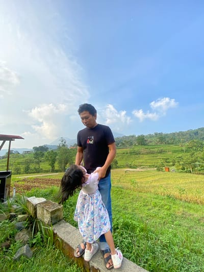 In Penanggungan, Indonesia, a man in a black shirt and blue jeans is holding a young girl in a floral dress. She is leaning back, with her head and hair dangling over the edge of a concrete barrier. The background includes rice paddies, green foliage, a hillside, and a blue sky with scattered clouds.