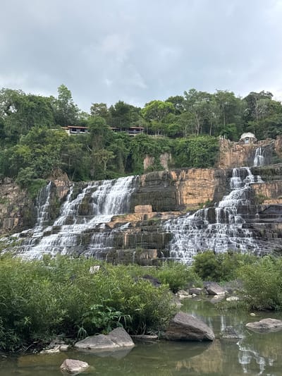The image depicts a wide, multi-tiered waterfall cascading down a rocky cliff face. Lush green vegetation, including trees and bushes, surrounds the waterfall and the cliff. Several buildings, appearing to be resort-style accommodations with balconies or verandas, are visible nestled among the trees at the top of the cliff. A small, ornate structure, possibly a pavilion or shrine, is also situated on the upper level. In the foreground, the water at the base of the waterfall is calm and reflects the greenery and the sky. Several large rocks are present in the shallow water. The sky is overcast, suggesting a cloudy or possibly rainy day. The overall scene conveys a natural landscape with developed tourist amenities. The location is identified as Datanla Waterfall in the city of Tân Phú, Vietnam.