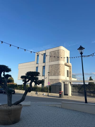 This is a vertical, eye-level, outdoor photograph taken in Chernomorets, Bulgaria. The image depicts a modern building with a light-colored facade and stone accents. In the foreground, there is a string of lights with exposed bulbs extending across the frame. A lamppost with a decorative fixture stands on the left side of the image, supporting the string of lights.
To the right of the building, a dark metal fence encloses the property. In front of the building and to the right of the fence, a paved walkway is visible. A large, twisted tree, possibly a bonsai or olive tree, is planted in a burlap-covered pot filled with pebbles. It is partially obscuring the building and the sidewalk. A street runs alongside the sidewalk, with a car parked on the left side of the street. A red sign with white text, potentially indicating a parking restriction or information, is visible near the fence. Another traffic sign, a red circle with a horizontal bar, is also present.
The sky is clear and blue, suggesting a sunny day. The sun is casting shadows, indicating it is likely daytime, possibly late morning or afternoon. The overall scene suggests a well-maintained urban or semi-urban environment, possibly a commercial or residential area with a focus on modern architecture and landscaping. The clear sky and bright sunlight create a cheerful and pleasant atmosphere. No people are visible in the frame.