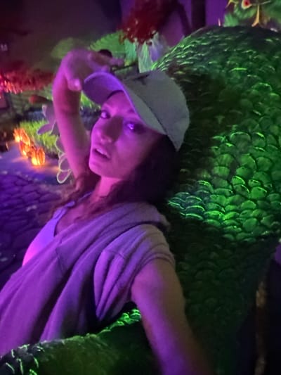 A woman in Binh Lu, Vietnam is leaning against a large, green, scaled structure that resembles a mythical creature or a themed display, possibly a dragon. She is wearing a baseball cap and a light-colored top with a purple hue from the lighting. The environment is dark with neon and artificial lighting, creating a surreal or fantasy-like atmosphere. There are artificial plants and decorations around, suggesting a tourist attraction or themed entertainment area.
