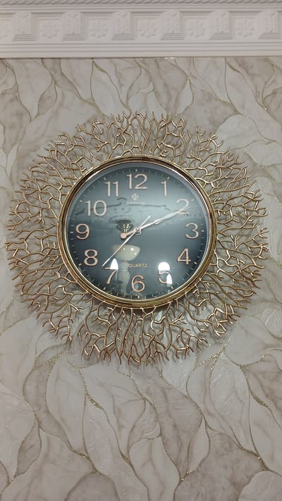 A close-up, eye-level, full shot captures a round, golden, decorative wall clock with a dark face and rose gold numerals, mounted on a beige and white marbled wallpaper. The clock features a branch-like, golden frame that extends outwards. The time on the clock reads approximately 12:06. Above the clock, a decorative white cornice molding is visible. The overall scene suggests an indoor setting, possibly a living room or hallway, in a residential building. The image does not provide information about the city of Yalama, Azerbaijan.