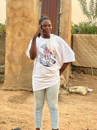 In Idiroko, Nigeria, an outdoor scene depicts a young woman standing on reddish-brown dirt ground in front of a light brown, textured wall. The woman has dark skin and hair pulled back, wearing a loose white t-shirt with a blue, red, and white "MONCLER" logo, and light-colored skinny jeans with split hems at the ankles. A white cloth is draped over her left shoulder. She holds a black smartphone to her right ear with her right hand, engaged in a call, and her left hand rests on her hip. Her gaze is directed slightly to her right. Behind her, a rusty corrugated metal door is visible on the brown wall. To the right, a light-colored, quilted mattress leans against a lower structure, with green foliage and sky in the background. A small, greyish-brown sack or package lies on the ground to the far right.