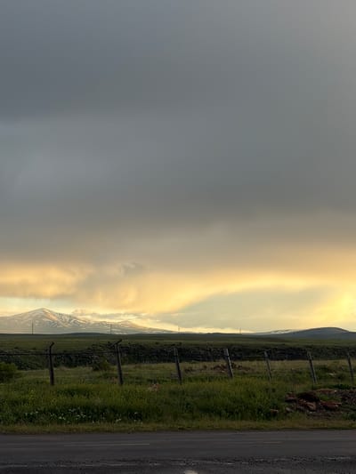 The image shows a landscape in Anipemza, Armenia. On the right side is a road with vegetation and a fence line. In the distance, mountains are visible, partially obscured by clouds and what appears to be a weather front or cloud formation. The sky is divided between a dark, overcast area on the left and a lighter, brighter area near the mountains.