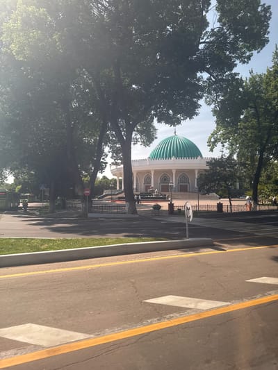 The image depicts a mosque with a prominent turquoise dome, situated in Kashgar mahalla, Uzbekistan. The mosque is surrounded by mature trees, partially obscuring the view. A street runs alongside the mosque with white and yellow lane markings. There are no people in the foreground. A fence surrounds the mosque. Signage is visible.