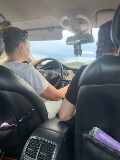The image is taken from the back seat of a car, looking forward. The driver, a woman with blonde hair pulled back in a ponytail and wearing sunglasses on her head, is visible from the shoulders up. She is wearing a light grey t-shirt and has her hands on the steering wheel, which has a Mercedes-Benz logo. Next to her, in the passenger seat, is another person, seen from the back of their head, with dark hair in braids. They are wearing a black t-shirt. A dashboard air freshener, shaped like a skull, hangs from the rearview mirror. The interior of the car appears to be black leather. Through the windshield, a cloudy sky and a green landscape with trees are visible. The location is specified as Курапольскі сельскі Савет, Belarus.
