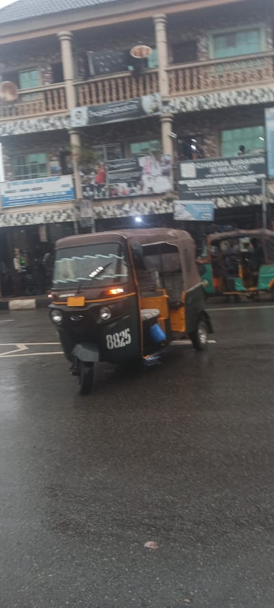 The image depicts a motorized tricycle, commonly known as a "keke", in Umuahia, Nigeria. The keke is primarily dark brown and yellow with the number "8825" displayed on its front. The vehicle is moving on a wet asphalt road. In the background is a two-story building with a detailed balcony. The building appears to house multiple businesses, indicated by the signs above the storefronts. The overcast sky suggests recent or ongoing rain. The scene appears to be a common urban setting in Umuahia.