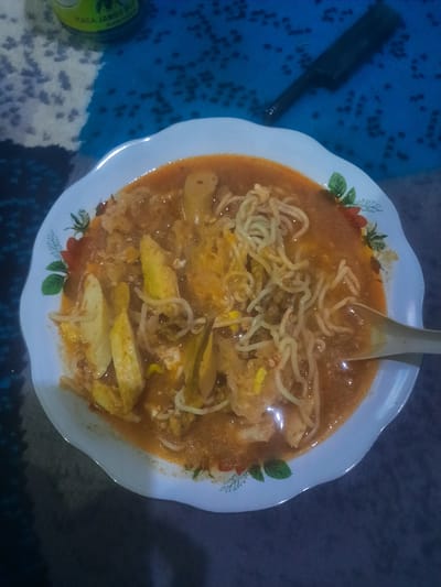 A close-up, overhead shot shows a plate of noodle soup on a patterned surface. The main subject is a white, scalloped-edge plate filled with a reddish-orange broth, thin yellow noodles, and pieces of what appear to be fried egg or omelet, along with some lighter colored vegetable strips, possibly bean sprouts or cabbage. A light-colored spoon is resting in the center of the dish. The rim of the plate is adorned with a floral design featuring red flowers and green leaves.
The plate rests on a blue and white patterned surface, likely a tablecloth or mat, which is visible in the foreground and extends into the background. To the left of the plate, partially out of frame, a dark, elongated object resembling a pen or marker lies on the blue patterned surface. In the bottom left corner, a portion of a green can is visible, with the white text "RASA JAMBU" printed vertically, suggesting it is a guava-flavored drink. The overall lighting appears dim, possibly indicating an indoor setting or evening time. The scene suggests a casual meal.