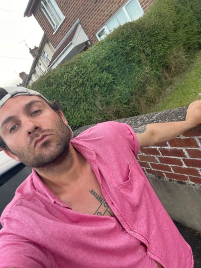 A man wearing a pink short-sleeved collared shirt and a grey baseball cap is leaning on a brick wall. He has dark hair, stubble, and blue eyes. He has tattoos on his chest and left arm. Behind him is a row of houses with green hedges and a grassy lawn. The scene appears to be in a suburban area of Moy, United Kingdom.