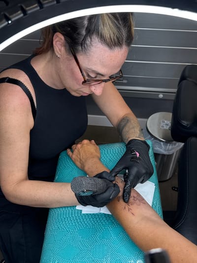 The image depicts a woman tattooing a floral design on a person's arm. The woman, wearing glasses, a black tank top, and with her hair pulled back, is focused on her work. She wears black gloves with a small pink decoration on the back of one hand. The tattoo is being applied to the inner forearm of a person whose arm rests on a turquoise snake-skin patterned surface. A white paper towel is visible underneath the arm. A circular light source is positioned to the left, and black chairs are above and behind. A metal stand is near the turquoise surface. No specific details in the image suggest the location is Yaiza, Spain.