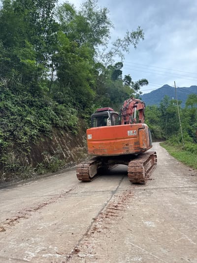 An orange excavator is stationary on a paved road in Cổ Lũng Commune, Vietnam. The road is wet and shows signs of mud or dirt. Lush green vegetation covers the steep embankment to the left of the road. In the background, a winding road leads towards rolling hills and mountains under a cloudy sky. Power lines are visible in the distance. The excavator appears to be the primary subject of the image, positioned centrally in the frame, facing away from the viewer and slightly to the right.