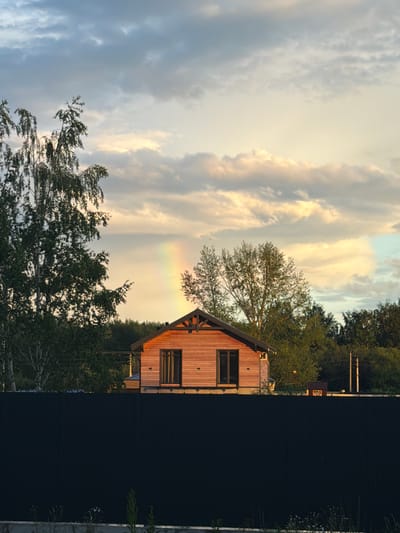 The image captures a natural scene with a modern wooden house against a dramatic sky. The house, constructed with vertical wooden planks, features a prominent gable roof with exposed beams and a small, dark-framed window on the upper floor. Its rustic design contrasts with the surrounding environment.

The sky is a dominant element, filled with a mix of light and dark clouds. The clouds are illuminated by the setting sun, casting a warm, golden glow on some parts of the sky, while other sections remain in shadow. A faint rainbow is visible in the sky, hinting at recent or ongoing rain.

In the foreground, the lush green canopy of trees, including some delicate, leafy branches, partially obscures the view of the house and sky. The trees suggest a wooded or semi-rural location. To the right of the house, a dark, solid fence runs vertically, creating a stark visual divide in the image. Beyond the fence and the house, more dense foliage and trees are visible, suggesting a natural setting.

The lighting indicates that it is either early morning or late afternoon, with the sun low on the horizon. The presence of clouds and a rainbow points to dynamic weather conditions. The overall impression is one of natural beauty and a peaceful, serene atmosphere, with the modern house subtly integrated into the landscape. No visible text is present in the image.