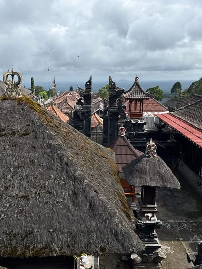 This image depicts a view of the Pura Lempuyang Luhur temple complex in Rendang, Indonesia, under a cloudy sky. The foreground is dominated by a large, weathered thatched roof, likely part of a pavilion or building. Beyond this, traditional Balinese architecture is visible, characterized by dark stone gate structures, tiled roofs in shades of red and brown, and tiered pagodas. Decorative elements, such as crown-like finials on some of the structures, are also present. In the background, a lush, hilly landscape stretches towards the horizon, with trees and distant mountains visible through the overcast conditions. The scene appears to be a serene and spiritual location. There are no people visible in the frame.