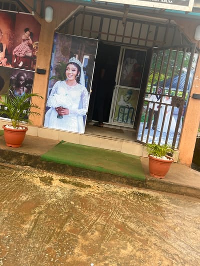 The image depicts the exterior of a business establishment, likely a photo studio, in Sabojibi, Nigeria. Prominently displayed are large posters of people, primarily featuring women in bridal attire and children. One poster shows a woman in a white wedding dress and tiara, holding a bouquet. Another displays a young girl in a red and white checkered dress sitting on a chair. A poster visible through a metal gate shows a couple in wedding attire.

The building has a storefront with a metal gate and large glass panels, which reflect the surroundings, including trees and the sky. Two potted plants, one on each side of the entrance, flank the display. The ground in front of the establishment is a wet, mottled surface, suggesting it may have recently rained. The overall scene is a typical street-level view of a commercial establishment in a Nigerian city, focusing on visual displays to attract customers.