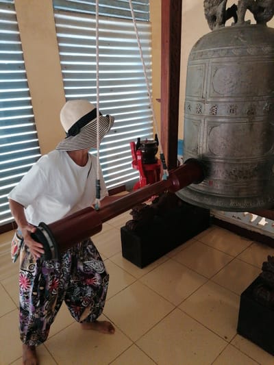 A person wearing a wide-brimmed hat and patterned pants is shown striking a large bell with a wooden mallet. The bell is ornate and appears to be made of bronze, with intricate carvings and inscriptions. The person is holding the mallet with both hands, poised to strike. The background features a wall with horizontal blinds, a wooden pillar, and a red structure with a black bowl-like object on top. The floor is tiled. The scene is likely within a temple or a similar cultural site.