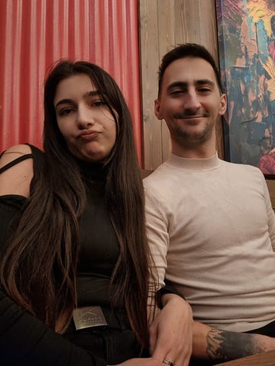 The image displays two individuals, a woman and a man, captured in a casual indoor setting, likely seated side-by-side. The image itself is oriented ninety degrees clockwise.
The woman is positioned on the left side of the frame (if viewed upright), with her face closer to the viewer. She has long, straight, dark brown hair that flows over her shoulders and chest. She is wearing a black long-sleeved top, which features a strap detail visible on her shoulder. Her expression is characterized by slightly pursed or "pouty" lips, with her dark eyes looking directly at the camera. A metallic, rectangular tag is visible on her black clothing near her shoulder, with the text "KUEBLER" written horizontally and "WORKWEAR" written vertically below it, alongside a stylized "K" logo. Her right hand is visible, resting on what appears to be the man's arm or lap.
The man is seated next to the woman, on the right side of the frame (if viewed upright). He has short, dark hair and light stubble on his face. He is wearing a light beige or off-white long-sleeved sweater with a ribbed crewneck collar. He has a gentle, closed-mouth smile, and is also looking towards the camera. On his left hand, a silver-colored ring is visible on his ring finger. His left forearm displays a distinctive tattoo, which depicts a landscape scene featuring mountains and trees, rendered in a black-and-grey shaded style that fades out towards his wrist.
The background behind the woman consists of a vibrant red wall with a distinct horizontal corrugated or paneled texture. Behind the man, there is a wooden wall made of horizontal planks, possibly stained a dark color. Below the man, a portion of a colorful, textured painting or artwork is visible, featuring abstract shapes and broad brushstrokes in shades of blue, yellow, and hints of red.
The overall mood appears relaxed and intimate, suggesting a shared moment between the two individuals.