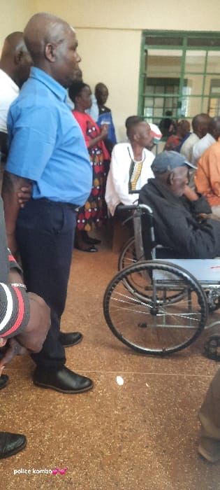 The scene depicts an indoor gathering within a building in Homa Bay, Kenya. The flooring is a light brown, speckled terrazzo-like material. The walls are plain, light beige. In the background, a window with green frames and horizontal security bars is visible.
In the immediate foreground on the left, an individual's lower arm, wearing a black and white striped wristband, is partially visible. Standing centrally in the foreground is a bald, dark-skinned male with a beard, wearing a light blue short-sleeved collared shirt, dark blue trousers, and black dress shoes. His hands are clasped behind his back, and he is facing right in profile.
To the right of the central standing figure, an elderly dark-skinned male is seated in a gray manual wheelchair. He wears a dark cap and a black long-sleeved top, with his head tilted slightly downward. His right hand is near his face. The wheelchair has silver spoked wheels with black tires.
Behind the central standing figure and the man in the wheelchair, a group of individuals is present. This includes a dark-skinned woman in a red patterned dress, several men, one wearing a white garment with dark patterned trim, and others partially obscured. Some individuals are standing, while others are seated in the background. No direct interactions between individuals are discernible; all appear to be in a waiting or observational state within the shared space.