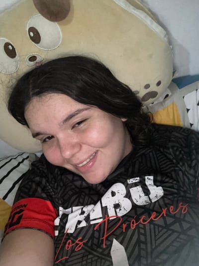 A young woman lies on her side on a bed, smiling slightly at the camera. Her head rests on a plush, light-brown animal pillow with darker brown spots. She is wearing a dark grey t-shirt with black geometric patterns and a white and red print of the word "Procesos". Red athletic shorts with dark lettering on the waistband peek out from beneath the t-shirt. The bed has a striped pillowcase and a patchwork quilt. The scene's location is unknown; there is no evidence suggesting San Juan de Los Morros, Venezuela.