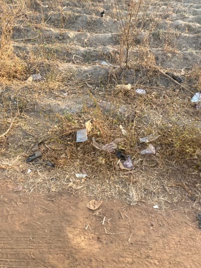 A view of ground with dry, sparse grass and weeds littered with discarded plastic and paper trash. The ground transitions to bare, reddish-brown soil on the right side, marked by faint tracks. The left portion features a more textured, compacted surface. The overall scene conveys a sense of neglect and environmental pollution.