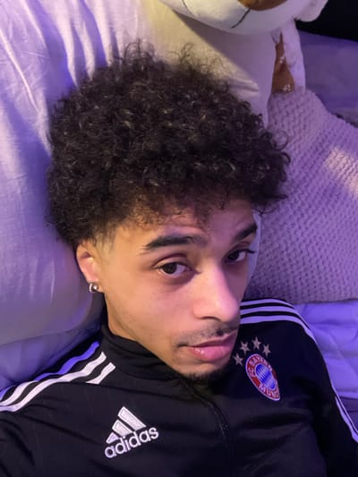The image depicts a young male subject positioned indoors, likely in a bed or lounge area, in the city of Plaisir, France. The individual has medium-brown skin, dark, tightly coiled curly hair, and dark eyes that are directed towards the viewer. He presents with faint facial hair, specifically a mustache and chin beard. A circular, silver-toned stud earring is visible in his left earlobe. He is attired in a black Adidas track jacket, featuring white stripes along the sleeves, the Adidas logo on the right side of the chest, and a circular "FC Bayern München" crest on the left side of the chest. The immediate background consists of soft furnishings, including a white pillow behind the subject's head, a textured light-colored pillow or blanket to the upper right, and a wrinkled, light purple sheet or blanket to the left. No specific actions beyond the subject's direct gaze and no other individuals or interactions are visible.