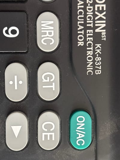 The image is a close-up, vertical shot of a portion of a black calculator. On the left, a large, black button displays the white numeral "9". To its right, a light grey, oval-shaped button has the white text "MRC" in all capital letters. Below "MRC" is another light grey, oval-shaped button with a white division symbol (÷). To the right of the division symbol is a light grey, oval button with the white letters "GT" in all capital letters. Below the "GT" button is another light grey, oval button with the white letters "CE" in all capital letters. To the left of the "CE" button is a light grey, oval button with a white downward-pointing triangle. In the bottom right of the image is a bright green, oval-shaped button with white text that reads "ON/AC" in all capital letters, oriented vertically. The calculator has a matte black surface that shows some dust or smudges. Text on the calculator's body, to the right of the buttons, is partially visible and appears to read "DEXIN BST" and "KK-837B" and "2-DIGIT ELECTRONIC CALCULATOR". The image does not contain any information about the location of Jrarat, Armenia.