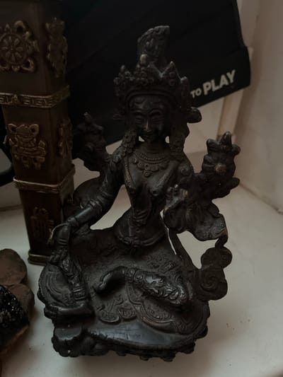 This is a close-up, slightly angled, indoor shot of a dark-colored, intricately detailed statue. The statue depicts a figure in motion, possibly dancing or in a dynamic pose, with multiple arms and elaborate headdress. The material appears to be metal, possibly bronze or brass, given its dark patina and the fine details that are visible. The statue is positioned on top of a decorative object, which has ornate carvings and metal embellishments, resembling a chest or a case.
In the background, to the left, a black object with white text is partially visible. The text reads "TO PLAY". The setting appears to be a dimly lit room, with a plain white wall as the backdrop. The lighting suggests it could be daytime, with natural light coming from an unseen source, casting shadows that highlight the texture of the statue and the decorative object. The overall mood is somewhat solemn and artistic, emphasizing the craftsmanship of the statue. There are no visible indications of weather or specific time of day beyond general indoor lighting.
The provided crops help to see the detailed facial features of the statue, the texture of its clothing, and the ornate designs on the supporting object. The last crop shows additional objects in the foreground, which appear to be natural materials like wood or stone, further adding to the eclectic nature of the displayed items. There are no explicit clues to the specific location beyond the mention of "Ukraine", and no discernible emotion is conveyed by the statue's features due to its stylized representation.