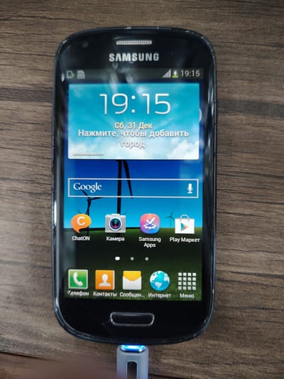 A Samsung smartphone is displayed on a wooden surface. The phone is black with a prominent screen showing the time as 19:15. Below the time, the screen displays weather information in Russian: "Сб, 31 Дек" (Sat, Dec 31) and "Нажмите, чтобы добавить город" (Press to add city). The Google search bar is visible, along with icons for ChatON, Camera, Samsung Apps, and Play Market. The bottom row of icons includes Phone, Contacts, Messages, Internet, and Menu. A blue light emanates from the charging port at the bottom of the phone. The wooden surface appears to be a table or desk. The context of Bokhtar, Tajikistan, is not evident from the image itself.