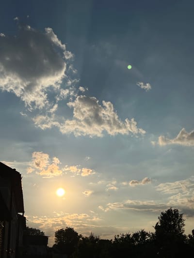 The image captures a sunset or sunrise scene in Ilavche, Ukraine. The sky is a gradient of blue and pale yellow, filled with scattered clouds. The sun, a bright orb, is visible in the lower left quadrant, casting a warm glow. In the upper portion of the sky, a small, bright green circular lens flare is present. Silhouetted against the sky are the rooftops of buildings and the tops of trees, suggesting a residential or semi-rural setting. The overall atmosphere is serene and bathed in soft light.