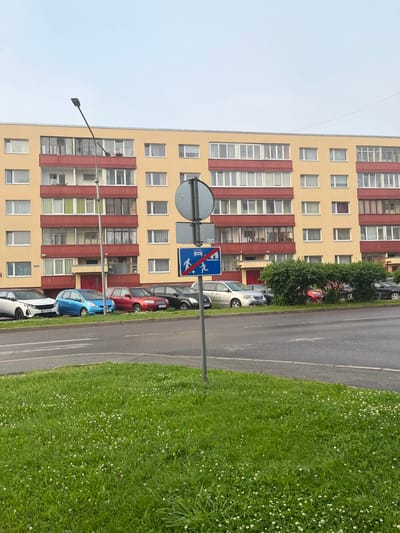 The image depicts a residential building in Maardu, Estonia. The building is a multi-story apartment complex with a yellow facade and red balconies. Several cars are parked in front of the building. A traffic sign indicating a pedestrian crossing and a no-entry symbol for vehicles is visible in the foreground. The area in front of the building is lined with green grass and small white flowers. The sky is overcast.