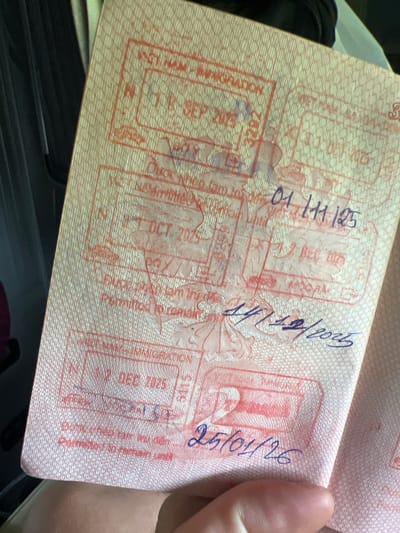 A passport page is angled with multiple entry/exit stamps and handwritten dates in blue ink. The stamps are in red ink and appear faded and partially obscured. The handwritten dates are "01/11/25", "11/12/2025" and "25/01/26". The stamp on the left contains "3282" and "SEP 2015", and below is "NA MIGRATION A". Another stamp contains "1 OCT 2015" and "Permitted to remain until". Another stamp contains "5115" and "11 DEC 2025" and "NA MIGRATION A". The location "Kon Ra Kiah, Vietnam" is not legible in this image. A human hand is partially visible holding the passport. The environment appears to be dimly lit.