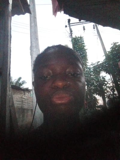 A person with dark skin is taking a selfie in what appears to be a residential area in Mbribit, Nigeria. Overhead power lines and poles are visible. Some vegetation is visible and there is a building made of wood.