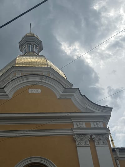 The image shows a close-up view of the dome and upper facade of a building, possibly a church, in Berezhany, Ukraine. The building is painted a pale yellow and features ornate white architectural details, including columns with Corinthian capitals and decorative friezes. A golden dome, textured with small diamond shapes, rises towards a cloudy, gray sky. The date "1900" is inscribed in a rectangular stone plaque on the facade. Dark power lines crisscross the sky, obscuring parts of the view. The overall impression is of a historic building under a dramatic sky.
