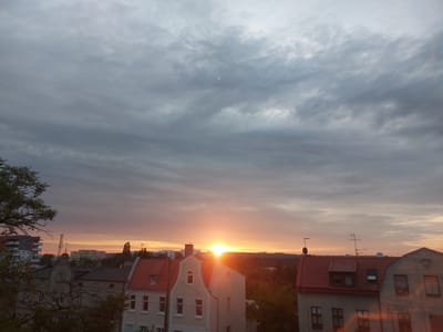 The image depicts a cityscape at sunset. The sky is filled with dramatic, overcast clouds in shades of gray and light purple. The sun, a bright orange orb, is low on the horizon, casting a warm glow across the scene. In the foreground, a row of buildings with red-tiled roofs is visible. These buildings appear to be residential, with visible windows and architectural details. Beyond these buildings, the landscape transitions to darker trees and the silhouettes of more distant structures. The overall impression is one of a tranquil, yet vibrant, evening in an urban setting. This scene is likely captured in Bydgoszcz, Poland, given the characteristic architecture and the presence of both older and more modern buildings.