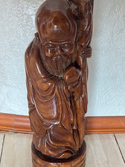 The image shows a wooden figurine of an elderly man with a long beard, dressed in flowing robes, holding a staff. The figurine is dark brown and highly polished, reflecting light from the environment. It stands on a circular wooden base. The background consists of a textured white wall and a wooden trim along the bottom. The lighting suggests an indoor setting. The specific location in Khostinskiy, Russia, cannot be determined from the visual information in the image.