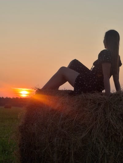 The image shows a silhouette of a young woman sitting on a haystack at sunset. She is wearing a dark dress with a floral pattern and is facing away from the camera, looking towards the horizon. The sun is setting, casting a warm orange glow across the sky and the surrounding field. In the background, a line of trees can be seen against the vibrant sunset. The haystack is large and appears to be in a rural setting, possibly on the outskirts of Perevoznoe, Russia. The overall mood of the image is peaceful and serene, with a hint of melancholy.