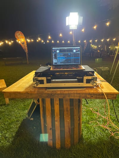 The image displays an outdoor DJ setup at night, suggesting an event taking place in Mururiini, Kenya. The scene is well-lit by various artificial light sources, creating a vibrant atmosphere.
In the center, a DJ station is positioned on a rustic, light-colored wooden table, possibly constructed from pallets. The main components of the DJ setup include a silver laptop, open and displaying a DJ software interface with visible waveforms and controls, and a DJ controller housed within a durable flight case. Text visible on the controller includes "Pioneer" and "Serato." Several wires and cables, including red and white ones, are connected to the equipment and extend across the table and onto the ground.
To the left of the DJ table, a tall stand holds a bright, square floodlight that powerfully illuminates the immediate area. In the background, numerous strings of warm, glowing Edison-style string lights crisscross the space, providing ambient lighting. Further back, a decorative, surfboard-shaped sign is visible, with the word "Guinness" partially discernible on it. The ground is covered in green grass, indicating an outdoor setting, possibly a garden or open field. The overall impression is that of an active outdoor party or gathering where music is being played after dark.