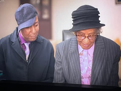 Two individuals are depicted, possibly engaged in a formal conversation or ceremony. The person on the left, a male, wears a dark jacket, a purple shirt, and a light purple tie. He is also wearing a dark blue cap that is tilted to the side. The person on the right, an older female, is wearing a dark pinstripe suit and a black ruffled hat. She has on glasses with tortoise-shell frames and a light pink shirt with a patterned design. Her gaze is directed downwards, while the male's gaze is also directed downwards and slightly to the right. The background is out of focus, suggesting an indoor setting, possibly a room with muted lighting and neutral-colored walls. There is no clear indication of a specific location within Liverpool, United Kingdom, or any specific objects or activities that would identify the scene as such.