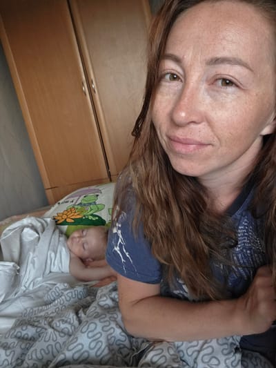 An adult female, appearing to be in her late 20s to early 30s, with auburn hair and a light complexion with freckles, is shown in a close-up selfie. She is wearing a blue t-shirt. She is in a room with a wooden wardrobe visible in the background. To her left, a baby is sleeping peacefully in a bed, covered with a white blanket and resting on a pillow with a frog illustration. The bedding has a gray wavy pattern. The overall scene suggests a domestic setting within a room in Буренка, Russia.
