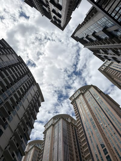 Looking up, four modern high-rise buildings surround the viewer, framing a sky filled with white clouds and patches of blue. The buildings, varying in design, are located in Taguig, Philippines. Each features rows of windows and balconies. There are no people visible.
