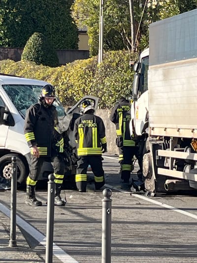 The scene depicts the aftermath of a two-vehicle collision on a paved road in Griante, Italy. A white van with a severely damaged front end and shattered windshield is positioned on the left, facing generally right. Adjacent to it, on the right, is a white truck with visible damage to its front section, specifically around the front wheel and bumper area.
Three uniformed Italian firefighters, identifiable by their dark protective suits with yellow reflective stripes and "VIGILI DEL FUOCO" text on their backs, are present. Each wears a dark helmet with yellow accents. One firefighter stands in the foreground, facing slightly left and looking down. A second firefighter is positioned centrally, facing away from the viewer, near the damaged van's front. The third firefighter is further right, also facing away, near the damaged front of the truck.
The road surface is asphalt, marked by a partial white lane stripe visible on the right. Several dark-colored bollards with spherical tops line the immediate foreground. The background features dense green foliage, including trees and bushes, suggesting a natural or semi-urban setting. The scene is illuminated by daylight, casting shadows on the road surface. Debris from the collision is visible on the road around the damaged vehicles.