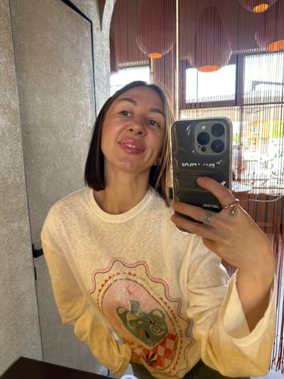 An adult female is depicted taking a mirror selfie in an interior setting located in Усады, Russia. She holds a dark-colored smartphone, consistent with an iPhone Pro Max model, encased in a clear protective cover, in her right hand. Reflected text "VWA" and "ЯJOЯ OT TОEА" is visible on the phone case. Her right hand displays multiple rings: a wide, multi-band ring on the ring finger and a ring with a charm (appearing as a clear bead and metal element) on the middle finger. She has medium-length dark brown hair, light complexion, and is smiling slightly with glossy pink lips, revealing upper teeth. A small gold-colored hoop earring is visible on her left ear. She is wearing a light-colored, long-sleeved t-shirt with a central graphic design featuring a scalloped border, a green anthropomorphic creature, checkerboard patterns, and floral elements in shades of pink, green, orange, and white. The background, reflected in the mirror, shows a textured light-colored wall on the left. To the right, a decorative screen of numerous reddish-brown vertical strings or beads is present. Behind this screen, multiple round, orange-glowing light fixtures are visible, along with large windows revealing a brightly lit outdoor environment with green foliage. A dark, flat surface occupies the immediate foreground.