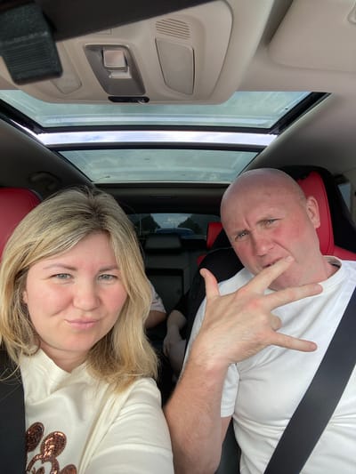 This is a selfie taken from inside a car, with the subjects positioned in the front seats. The image is oriented vertically. On the left, a woman with blonde hair is smiling at the camera. She is wearing a light-colored t-shirt decorated with sequined Mickey Mouse heads. On the right, a bald man with blue eyes is making a "rock on" hand gesture. He is wearing a white t-shirt and appears to be smiling or smirking.
The interior of the car features red and black seating, a black seatbelt, and a panoramic sunroof that reveals an overcast sky. The lighting suggests it is daytime. The scene appears to be set in a rural or suburban area, possibly in or near Новая Казмаска, Russia, given the context. The overall mood is casual and friendly. There is no visible text in the image.