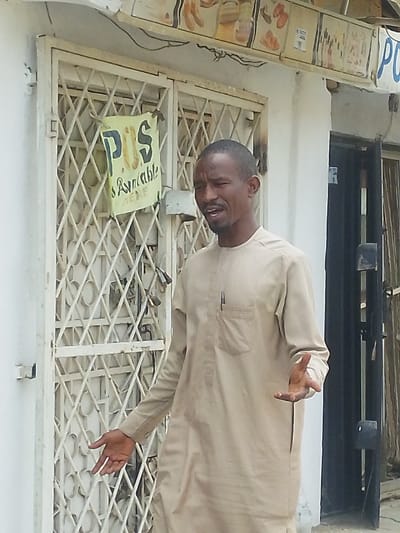 An adult male stands outdoors in front of a commercial building in Babban Giji, Nigeria, under daylight conditions. The man, with short dark hair and facial stubble, wears a light beige long-sleeved tunic with a chest pocket containing a black pen. He is captured with an open mouth and both hands gesturing, palms slightly open and facing upwards, indicating speech or explanation. Behind him is a white-walled building featuring a light-colored metal security gate with a diamond lattice pattern. A yellow, handmade sign reading "POS Available HERE" in dark lettering is affixed to the gate. Above the gate, a horizontal array of several rectangular signs or advertisements, some depicting food items, are visible. To the right of the man, a dark, recessed doorway is partially visible.