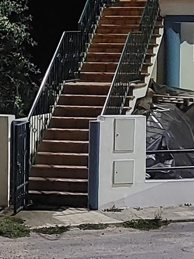 This image shows an outdoor staircase made of stone steps with a dark metal railing. The staircase is partially illuminated, creating shadows on the steps. To the left of the stairs, there is a dark green tree with leaves against a black background. To the right of the stairs, there is a light-colored building with two square electrical or utility boxes mounted on its exterior. Behind the utility boxes and to the right of the stairs, there is a pile of materials covered with a black tarp. The ground in front of the stairs is paved with concrete and asphalt, with patches of grass and weeds growing in the crevices. The scene appears to be a residential or commercial exterior. The location is identified as São Teotónio, Portugal.