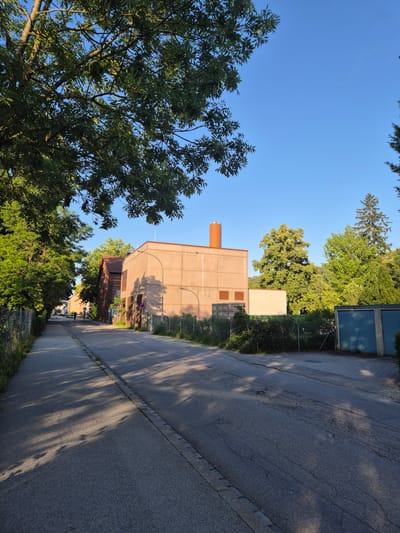 The image depicts a street scene in the city of Landshut, Germany, likely during daytime given the clear blue sky and the presence of shadows cast by trees onto the asphalt road. The road appears to be paved and shows some signs of wear, with cracks and unevenness visible in places.
On the left side of the road, a line of trees with lush green foliage creates a natural canopy, with branches extending over the street. Adjacent to the trees is a chain-link fence, beyond which is overgrown vegetation and a further fence. A red brick building with windows is visible further down the street on the left, with parked cars in front of it.
On the right side of the road, a light-brown, boxy industrial-style building dominates the foreground. This building has a rectangular orange chimney or ventilation shaft protruding from its roof. A smaller, lighter-colored structure is attached to the side of this building. A chain-link fence runs along the front of these buildings. Further to the right, a blue-colored corrugated metal structure, possibly a garage or storage unit, is visible.
The overall impression is of an industrial or commercial area on the outskirts of a city, with a mix of vegetation and buildings. The lighting suggests it is either morning or late afternoon, with the sun casting long shadows. There is one person visible in the distance on the left side of the road, appearing to be walking or cycling. No other people or significant actions are depicted.
