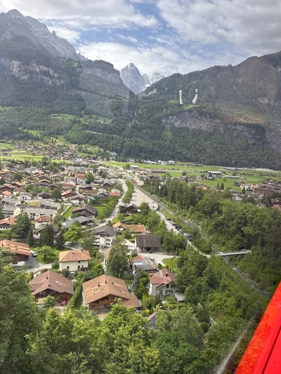 The image captures a panoramic view of a Swiss town nestled in a valley surrounded by lush green mountains. The architecture of the town is characterized by traditional Swiss chalets with steep, gabled roofs made of wood and tile. The houses are spread out, with many featuring well-maintained gardens and visible driveways. A river winds its way through the town, crossed by several small bridges, with a road running alongside it, lined with parked cars. The mountains in the background are heavily forested, with some rocky outcrops and snow-capped peaks visible in the distance under a cloudy sky. On the mountainside, two large, white structures resembling statues or pillars are visible. The overall impression is one of a peaceful, picturesque alpine village.