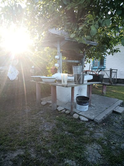 In Veremia, Ukraine, a well structure stands in a yard with a white house in the background. The sun shines brightly through the leaves of a large tree, creating lens flare. On a wooden table beneath the well structure, two large metal basins are present. One of these basins is filled with water. Also on the table are a tall glass pitcher, two metal buckets, and a lit candle. A large black plastic bucket with a white label is on the ground beside the table. The ground is covered with grass and patches of dirt.